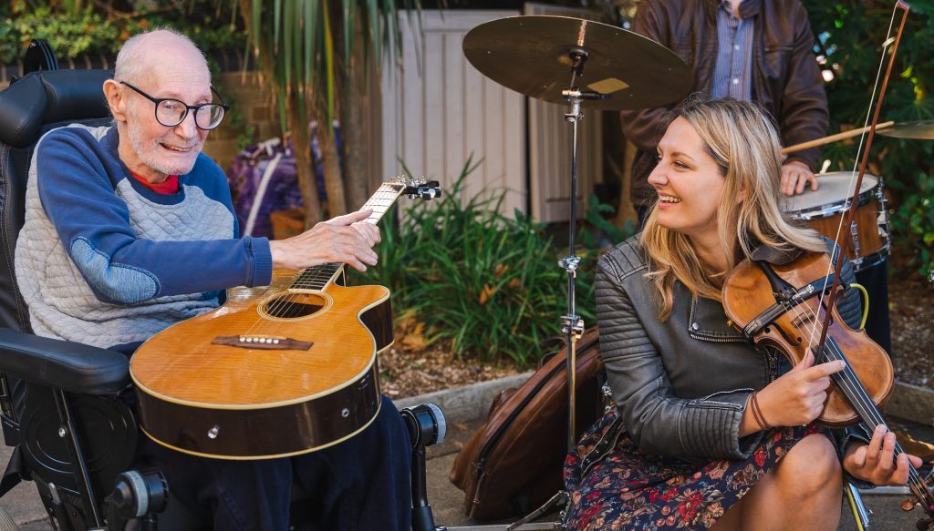 Resident John in a wheelchair outdoors holding an acoustic guitar watched by Alice Zawadzki holding a violin